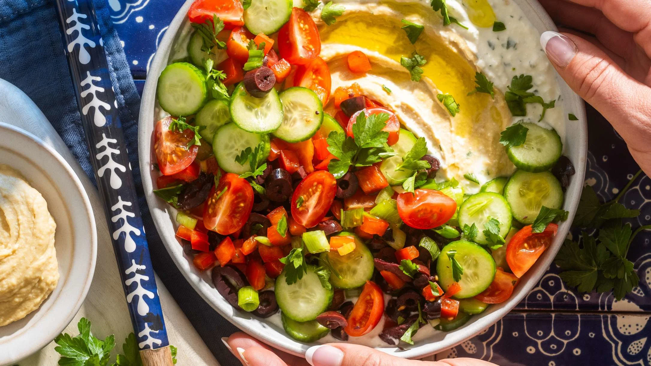 Mediterranean dip bowl with hummus topped with fresh cucumbers, tomatoes, olives, and herbs on a blue patterned cloth.