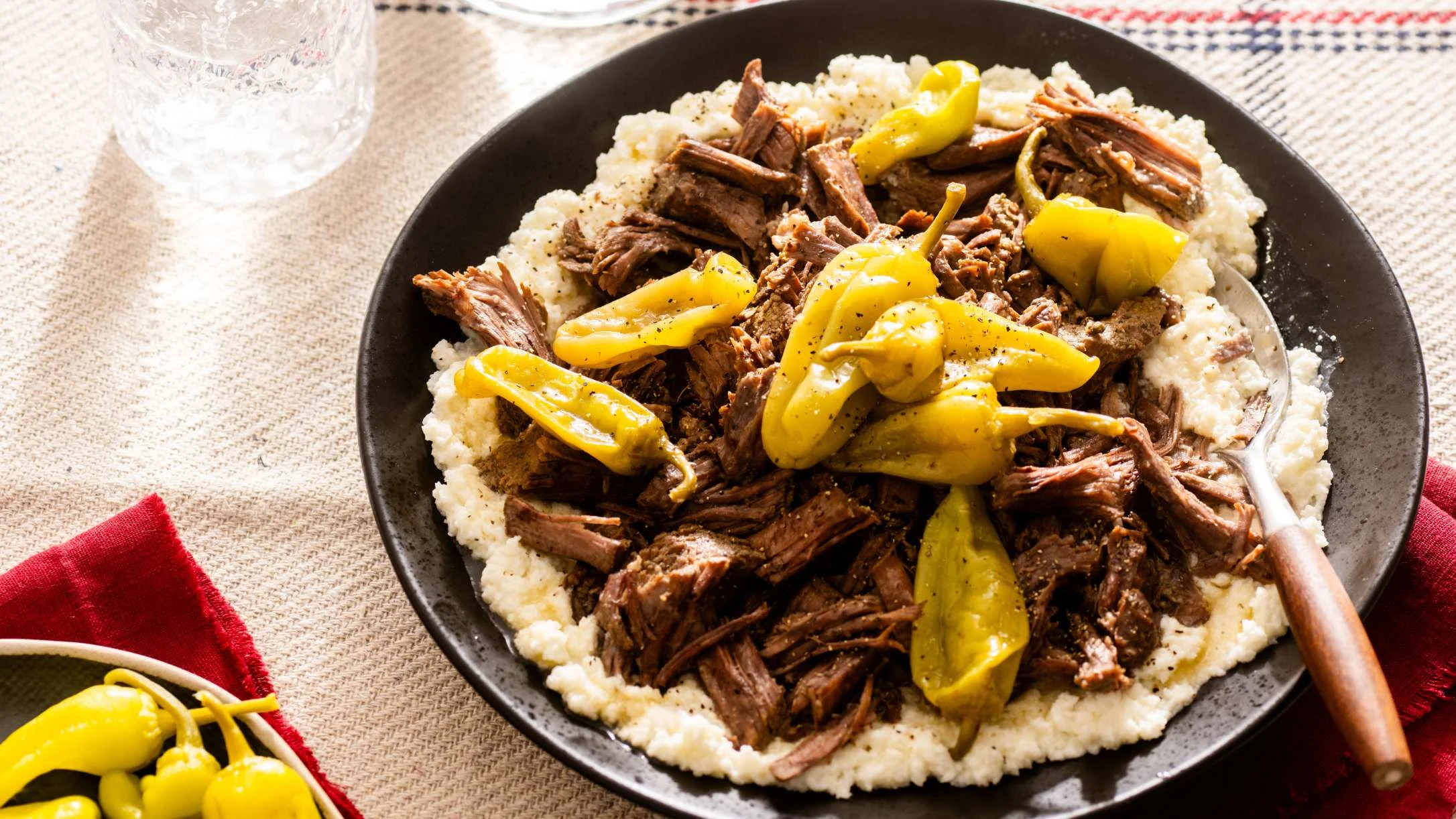 A dark bowl filled with creamy mashed cauliflower topped with shredded beef and yellow pepperoncini peppers, served with a spoon.