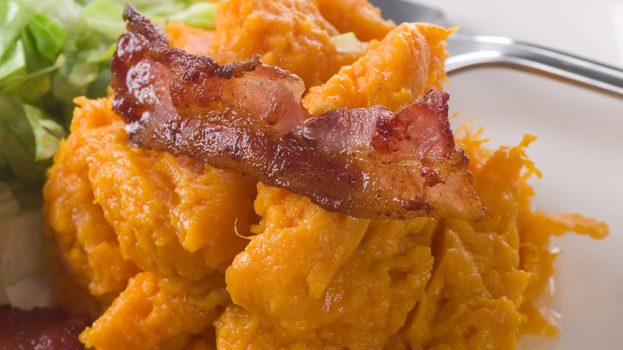 Mashed sweet potatoes topped with crispy bacon strips on a white plate, with fresh green lettuce in the background.