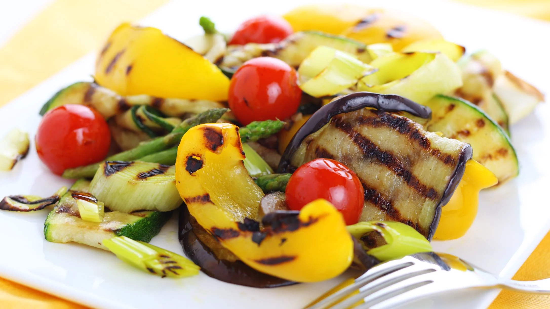 A white plate of colorful grilled vegetables including eggplant, zucchini, yellow peppers, cherry tomatoes, and asparagus, with a fork.