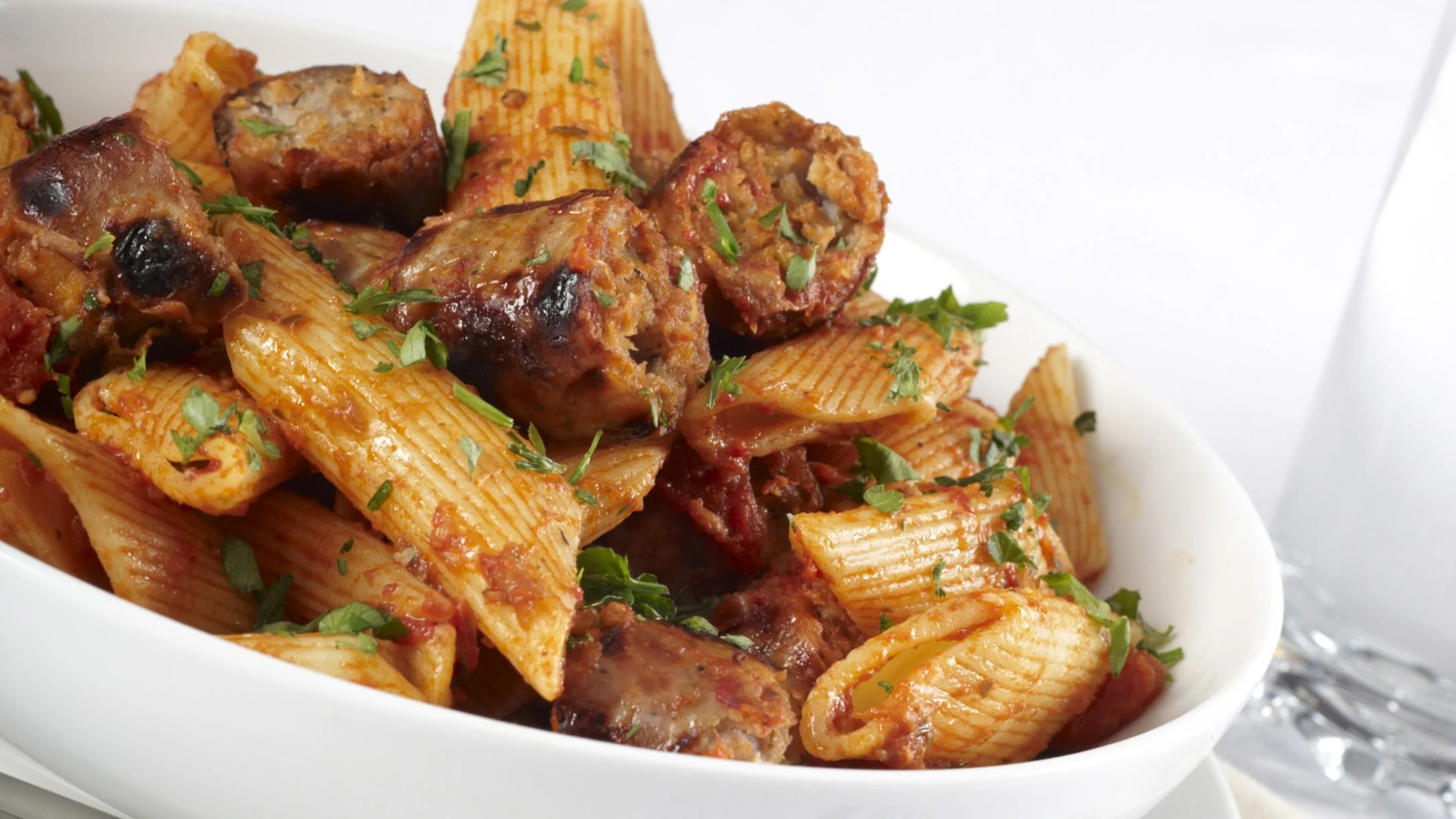 A white bowl filled with penne pasta and sliced sausage in tomato sauce, garnished with fresh chopped parsley.