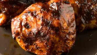 Closeup of glazed, caramelized BBQ chicken thighs with a rich, dark sauce and charred edges on a dark surface.