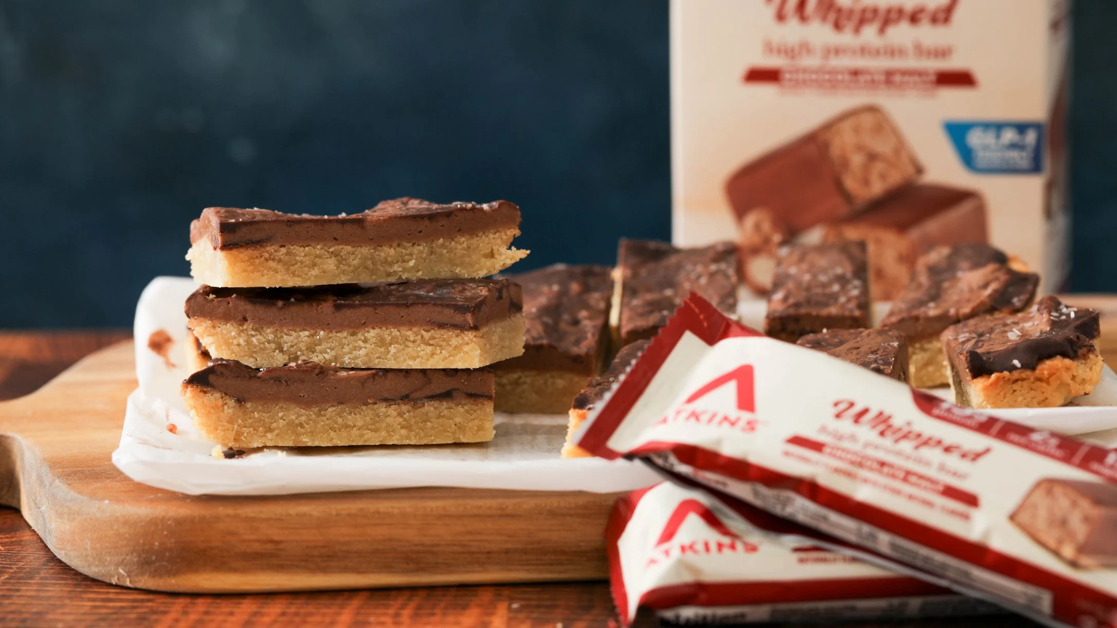 Stacked chocolate-topped cookie bars on a wooden board beside Atkins Whipped high protein bar wrappers and box.
