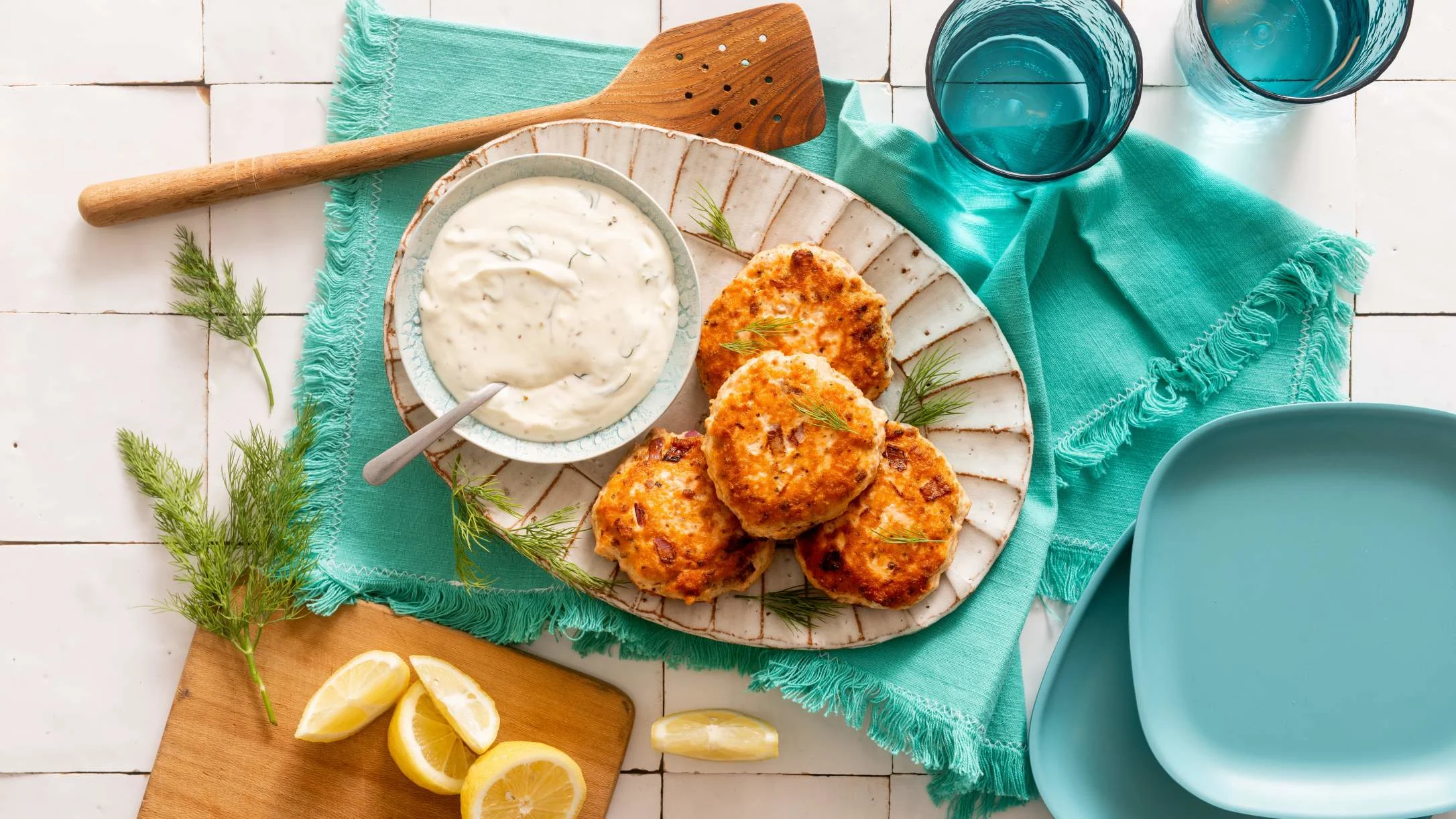 Golden fish cakes on a rustic plate with creamy dipping sauce, fresh dill, lemon wedges, and teal napkins.