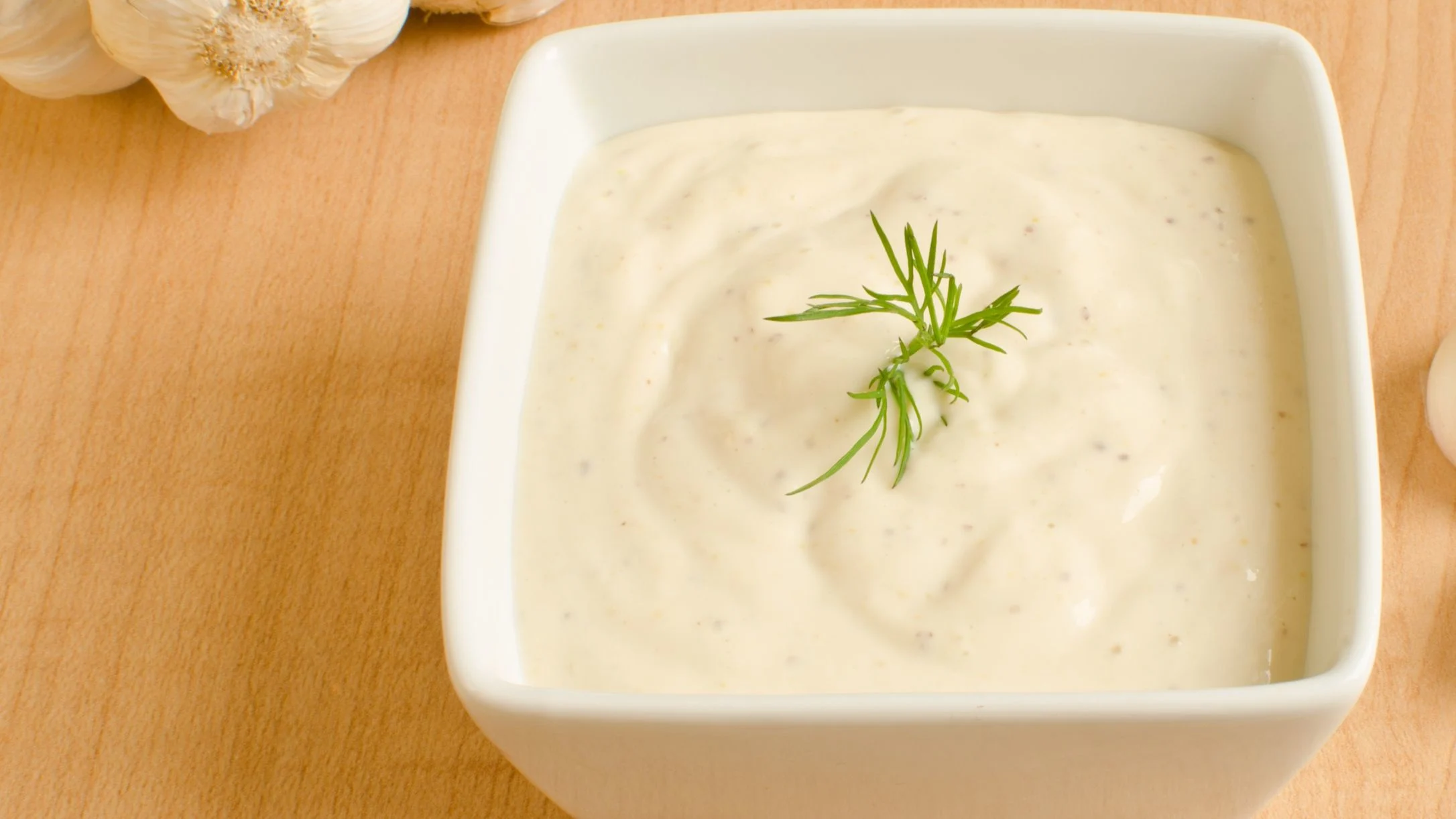 A white square bowl filled with creamy alfredo sauce, garnished with fresh herbs, placed on a wooden surface with garlic bulbs nearby.