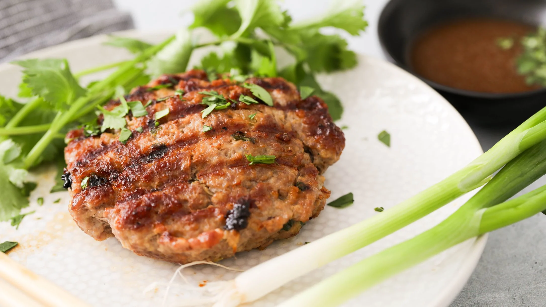 Grilled meat patty topped with fresh cilantro on a white plate, with green onions and a small bowl of dipping sauce.