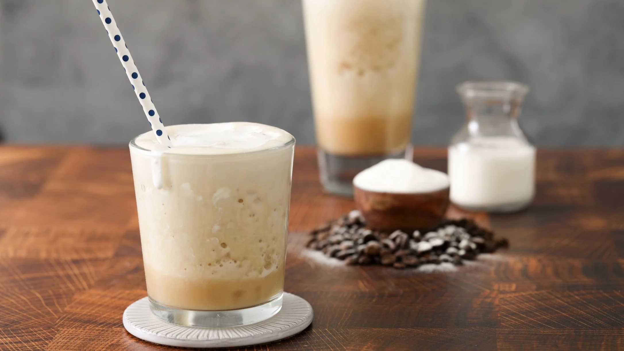 Frothy iced coffee drink with a polka-dot straw on a wooden table, surrounded by coffee beans, sugar, and milk.