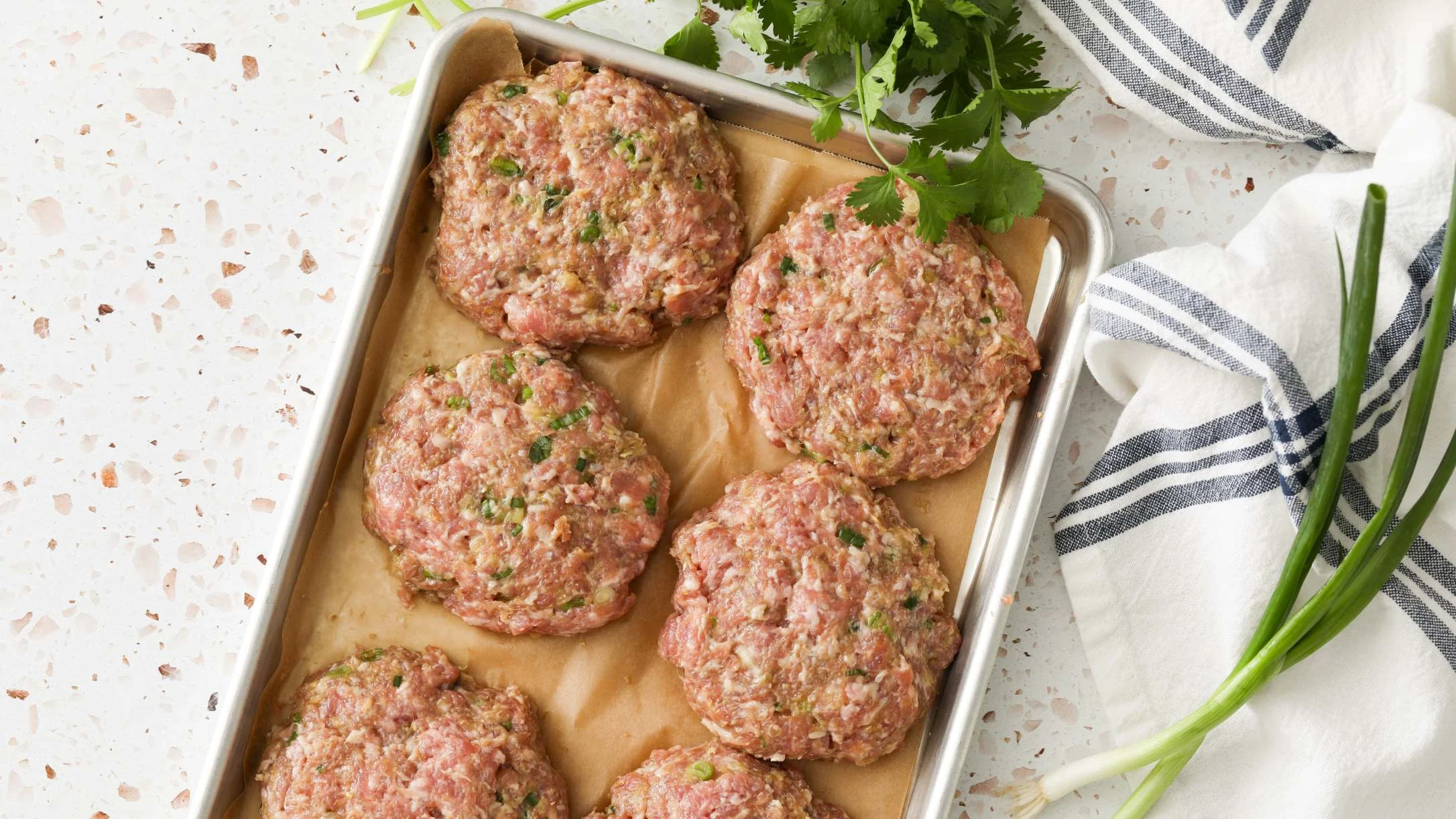 Six raw meat patties with herbs on a parchment-lined baking sheet, with fresh cilantro and green onions nearby.