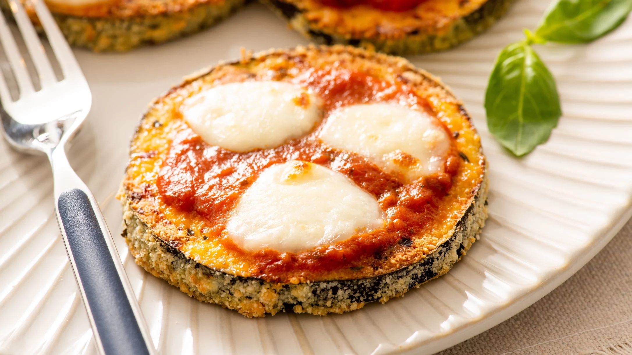 Breaded eggplant round topped with tomato sauce and melted mozzarella on a white plate, with fresh basil and a fork.