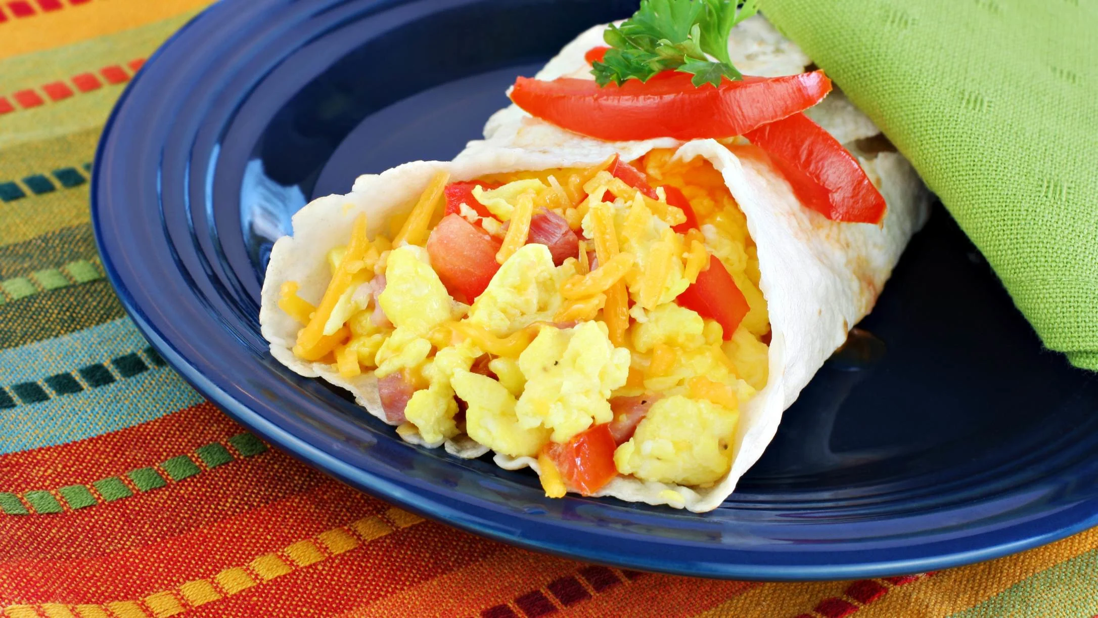 Breakfast burrito with scrambled eggs, cheese and tomatoes on a blue plate, garnished with fresh parsley and red pepper.