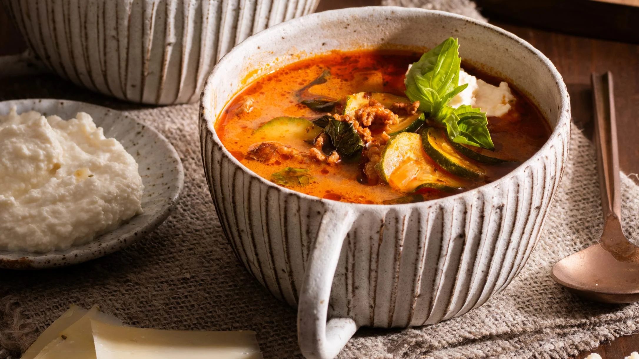 A rustic ceramic bowl of tomato-based soup with zucchini, meat, and fresh basil, served with ricotta cheese on the side.