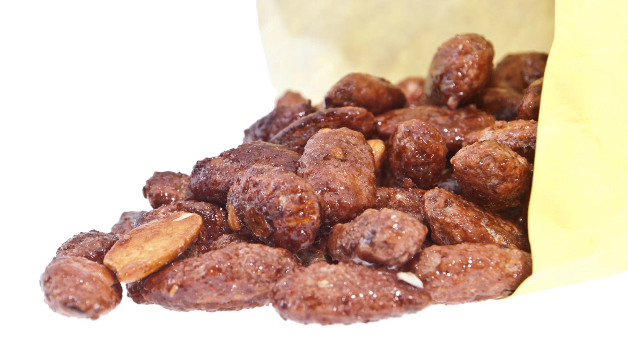 Candied nuts spilling out of an open kraft paper bag onto a white surface, coated in a shiny brown sugar glaze.