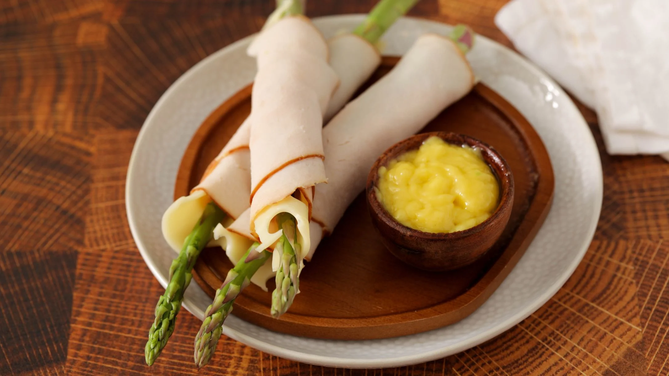 Asparagus spears wrapped in deli turkey and cheese on a plate, served with a small bowl of yellow dipping sauce.