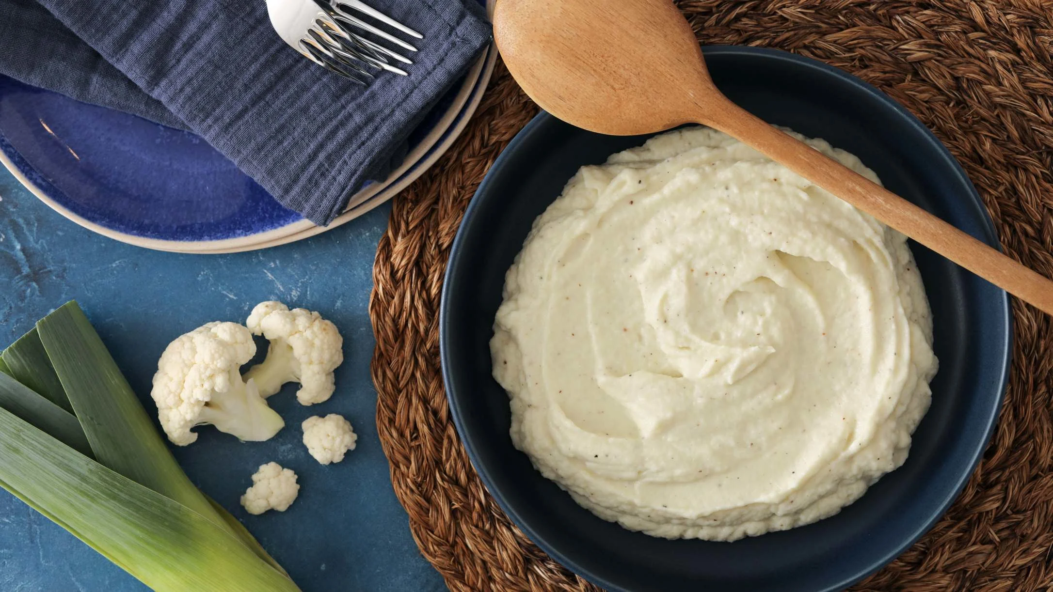 Puree of cauliflower in a blue bowl with a wood spoon, blue plates, cauliflower and leek on a blue background.