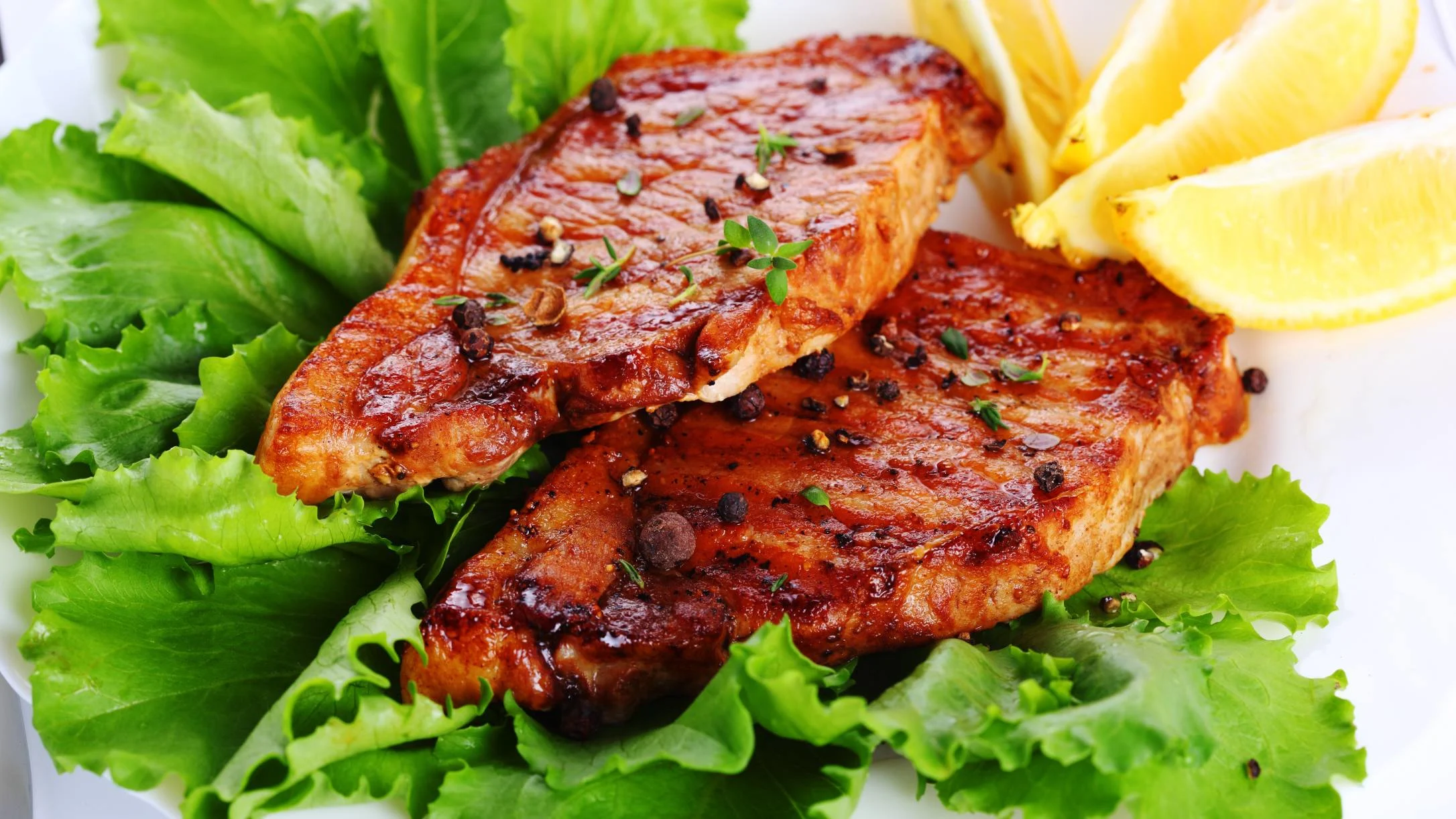 Two grilled pork chops seasoned with black pepper and herbs, served on fresh lettuce leaves with lemon wedges.