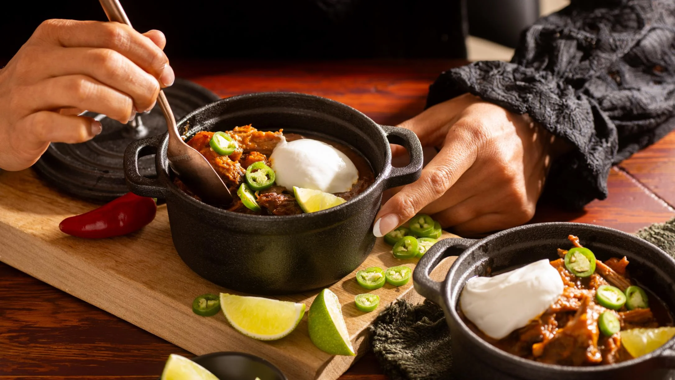 Hands serving chili in black cast iron mini pots topped with sour cream, jalapeños, and lime wedges on a wooden board.