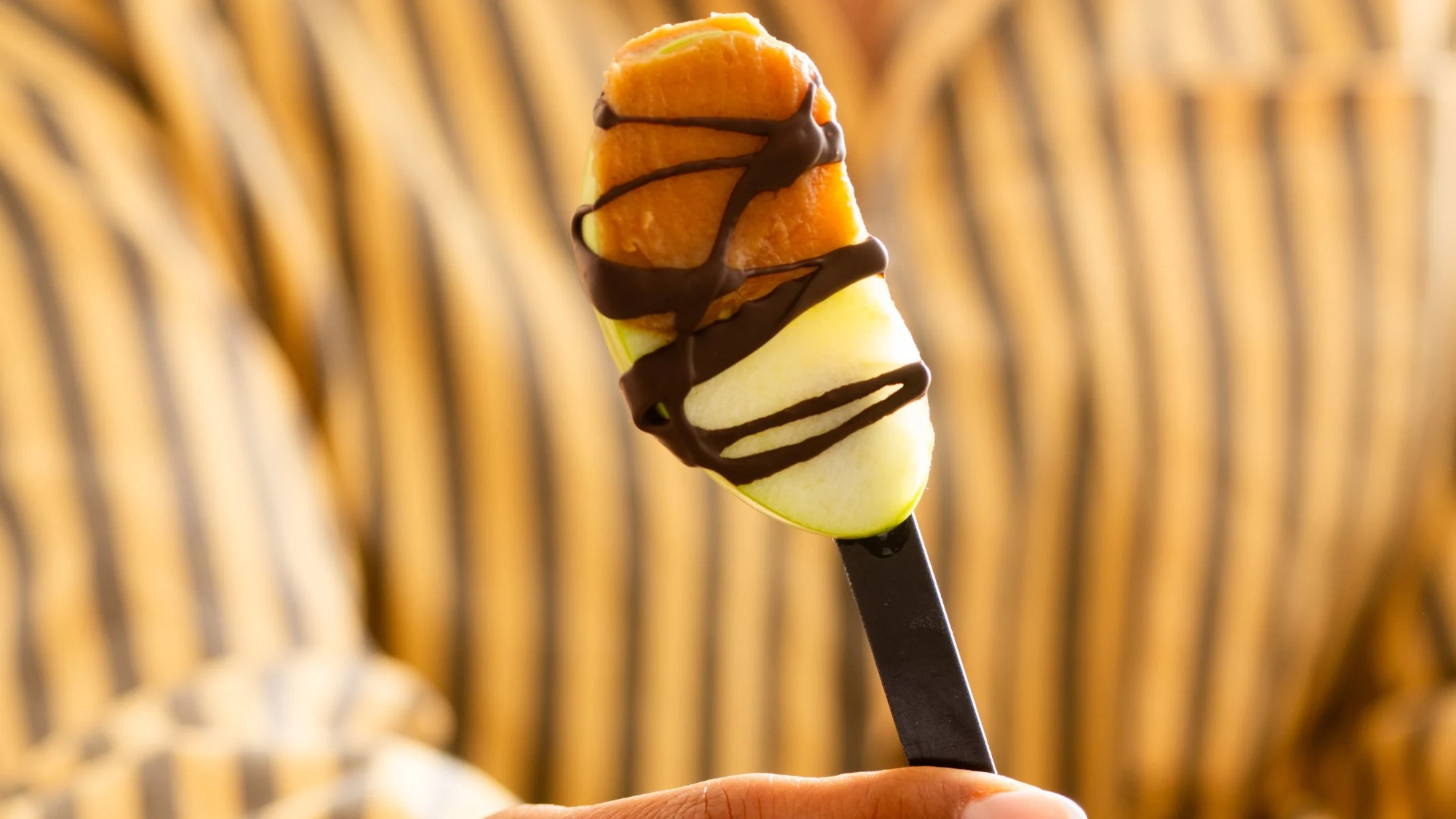 A hand holds a stick with an apple slice drizzled with dark chocolate, against a blurred striped background.