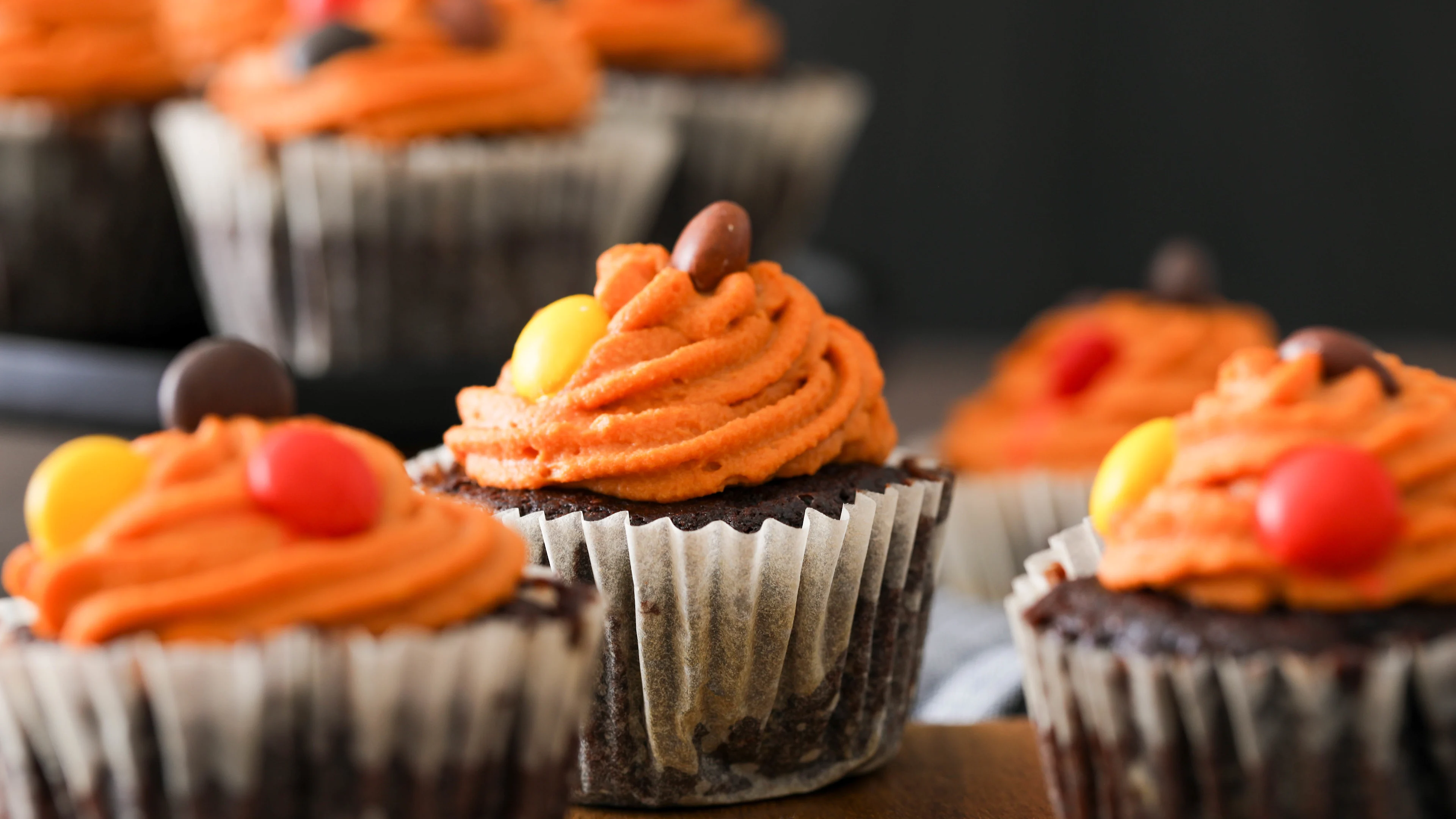 Chocolate cupcakes topped with swirled orange frosting and colorful candy pieces, displayed against a dark background.