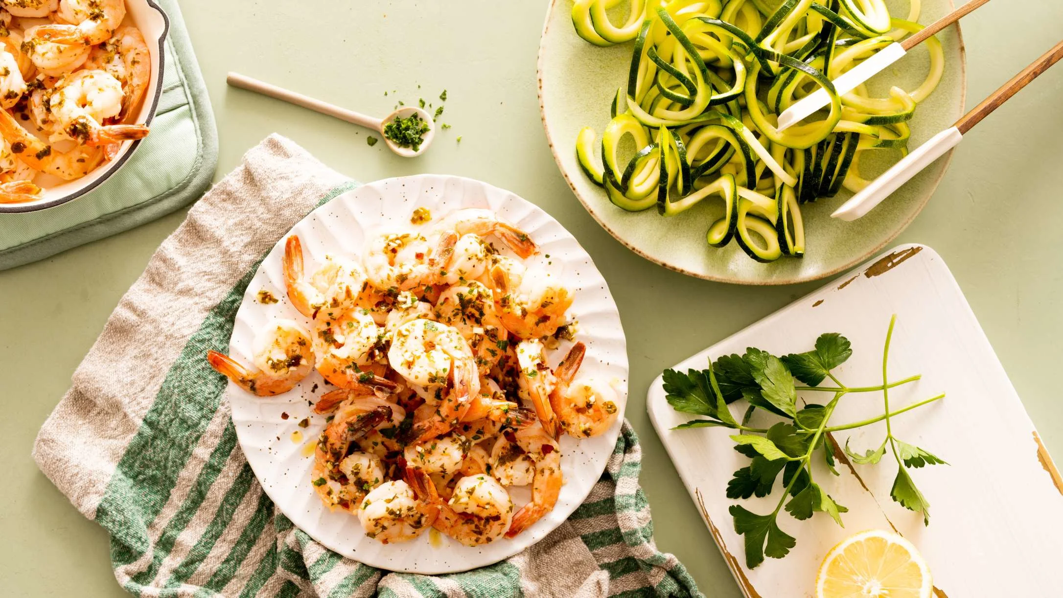 Herbed shrimp on a white plate beside a bowl of zucchini noodles, fresh parsley, lemon, and a baking dish of shrimp.