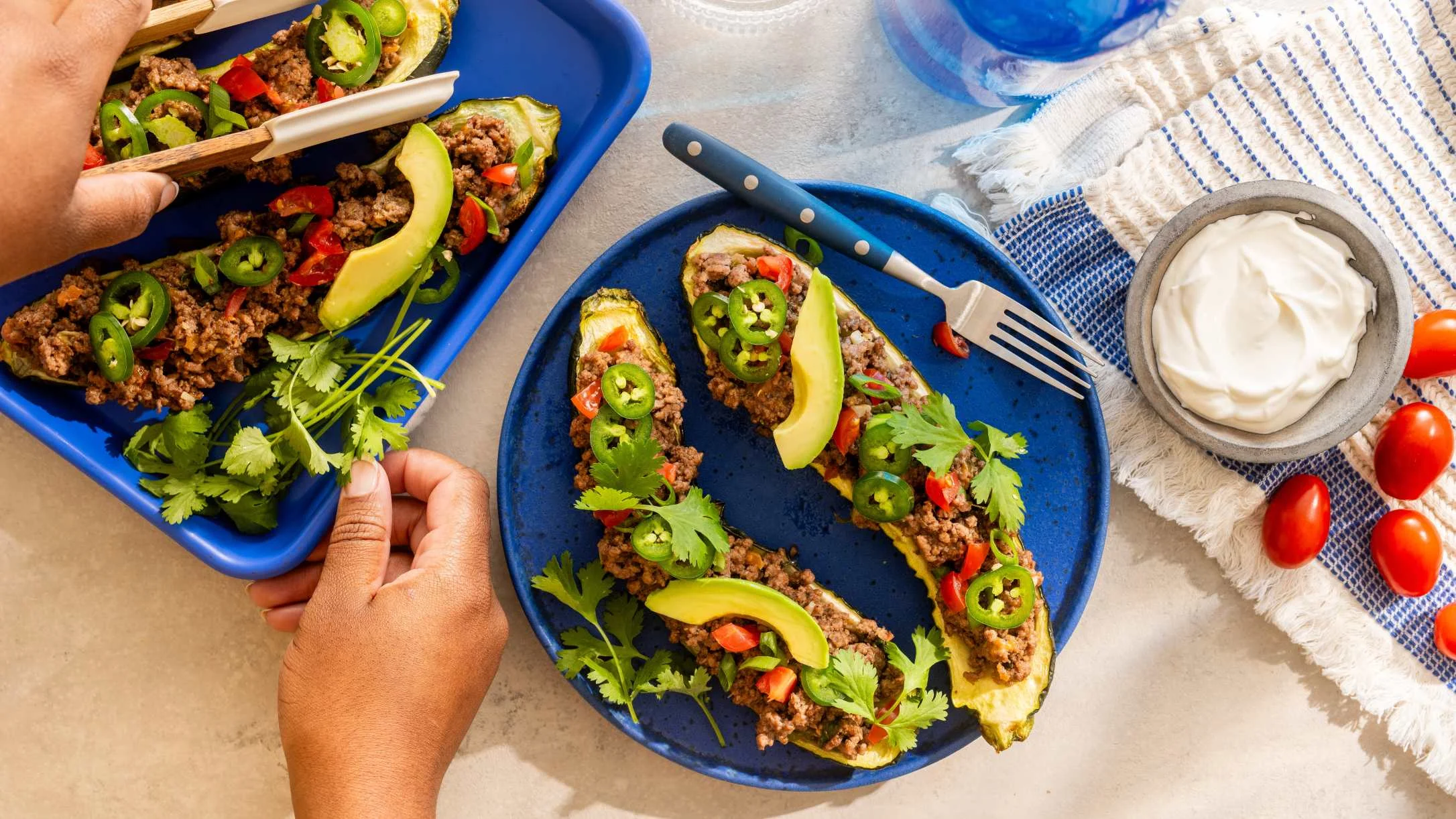 Stuffed zucchini boats filled with ground meat, jalapeños, avocado, and cilantro on blue plates, with sour cream nearby.