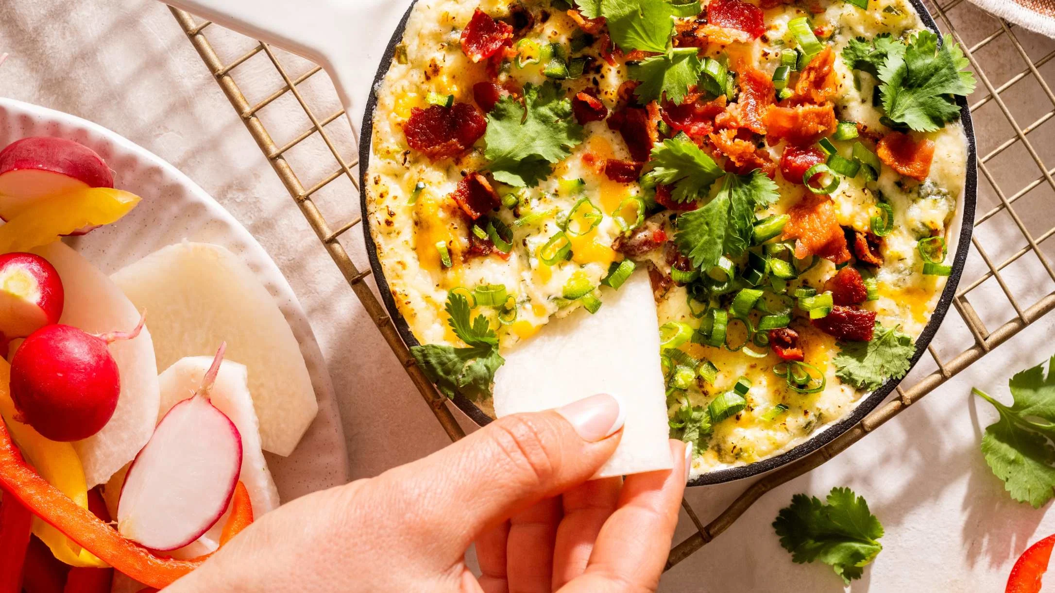 A hand dips a jicama slice into a cheesy skillet dip topped with bacon, cilantro, and green onions, beside colorful veggies.