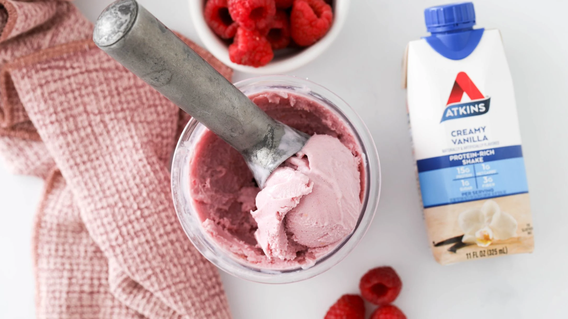 Pink raspberry ice cream being scooped, beside an Atkins Creamy Vanilla protein shake carton and fresh raspberries.