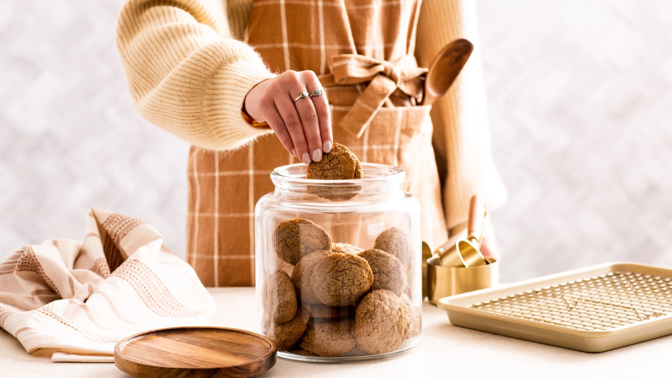 Person in cream sweater and tan apron taking a cookie from glass jar, with baking tray nearby.