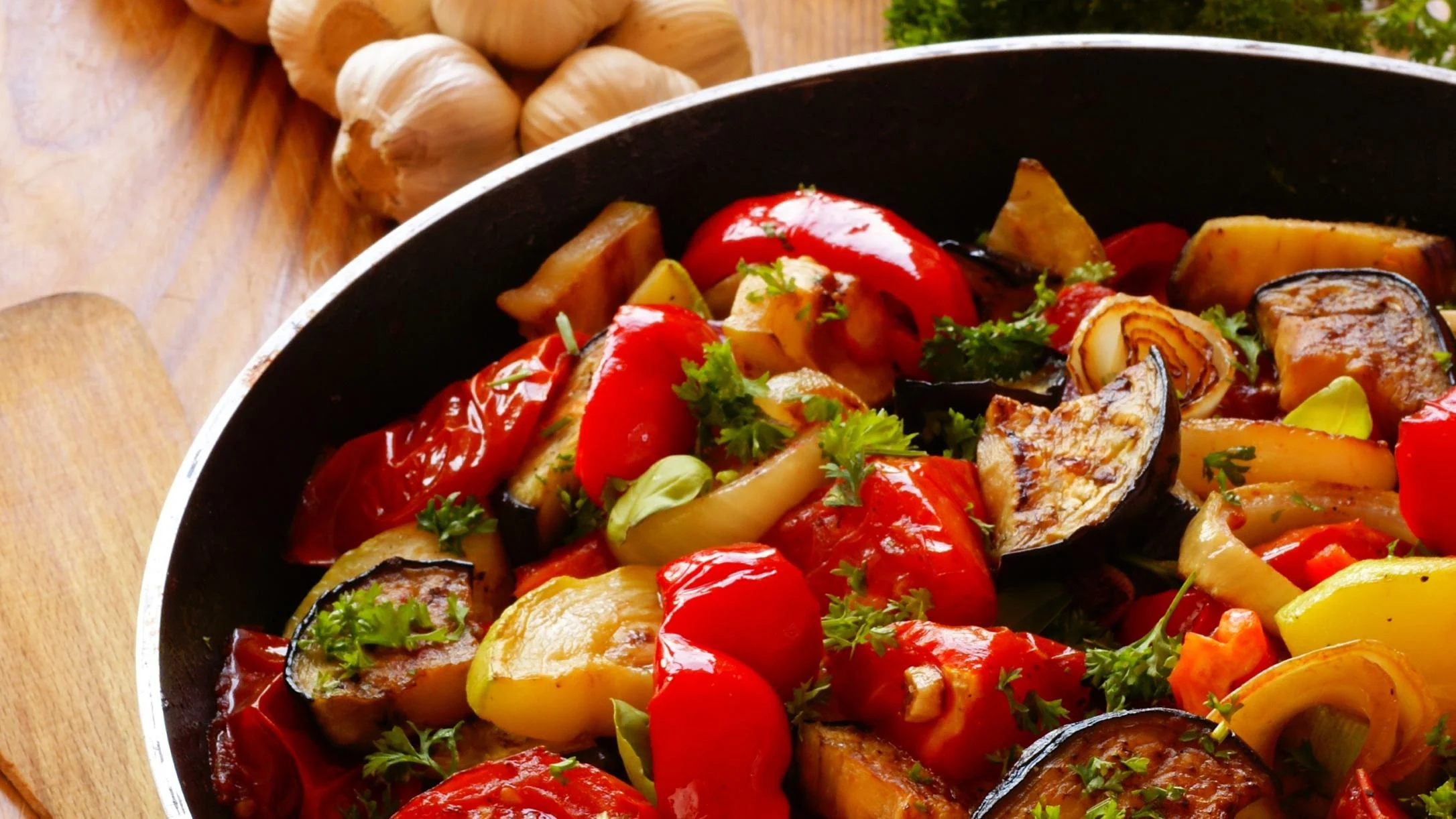 Colorful roasted vegetables in a black pan with red peppers, eggplant, and herbs, with garlic cloves in background.