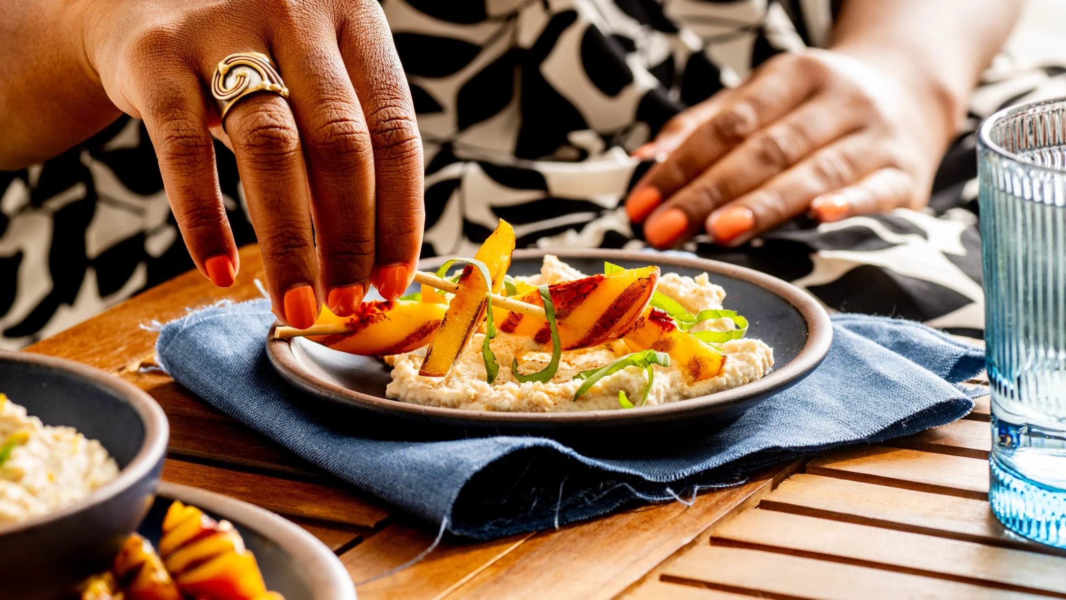 A woman with orange nails places peach slices on a creamy dish garnished with fresh basil, served in a dark bowl.