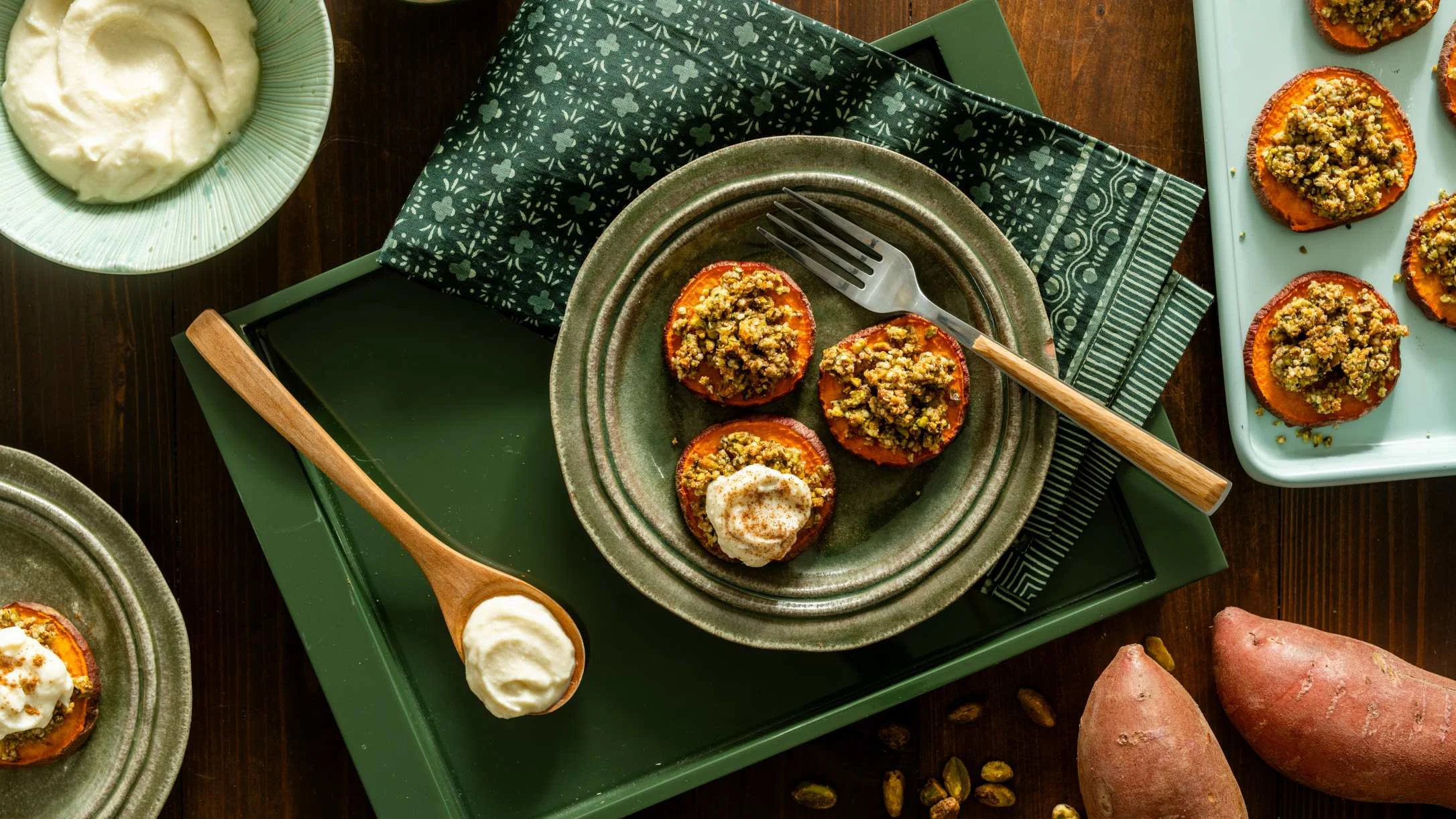 Roasted sweet potato rounds topped with pistachio crumble and cream on a green ceramic plate with a fork.