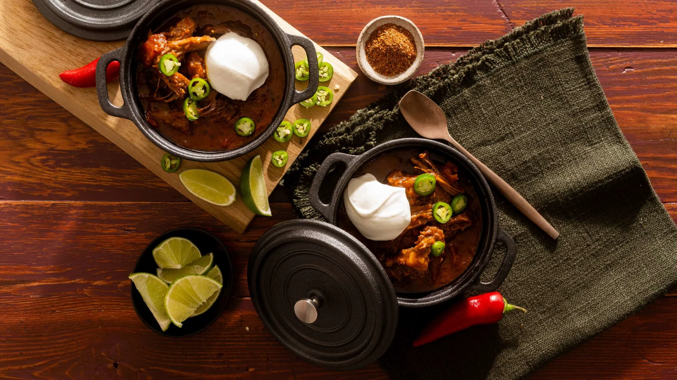 Two black cast iron pots of spicy meat stew topped with sour cream and jalapeños, served with lime wedges and spices.