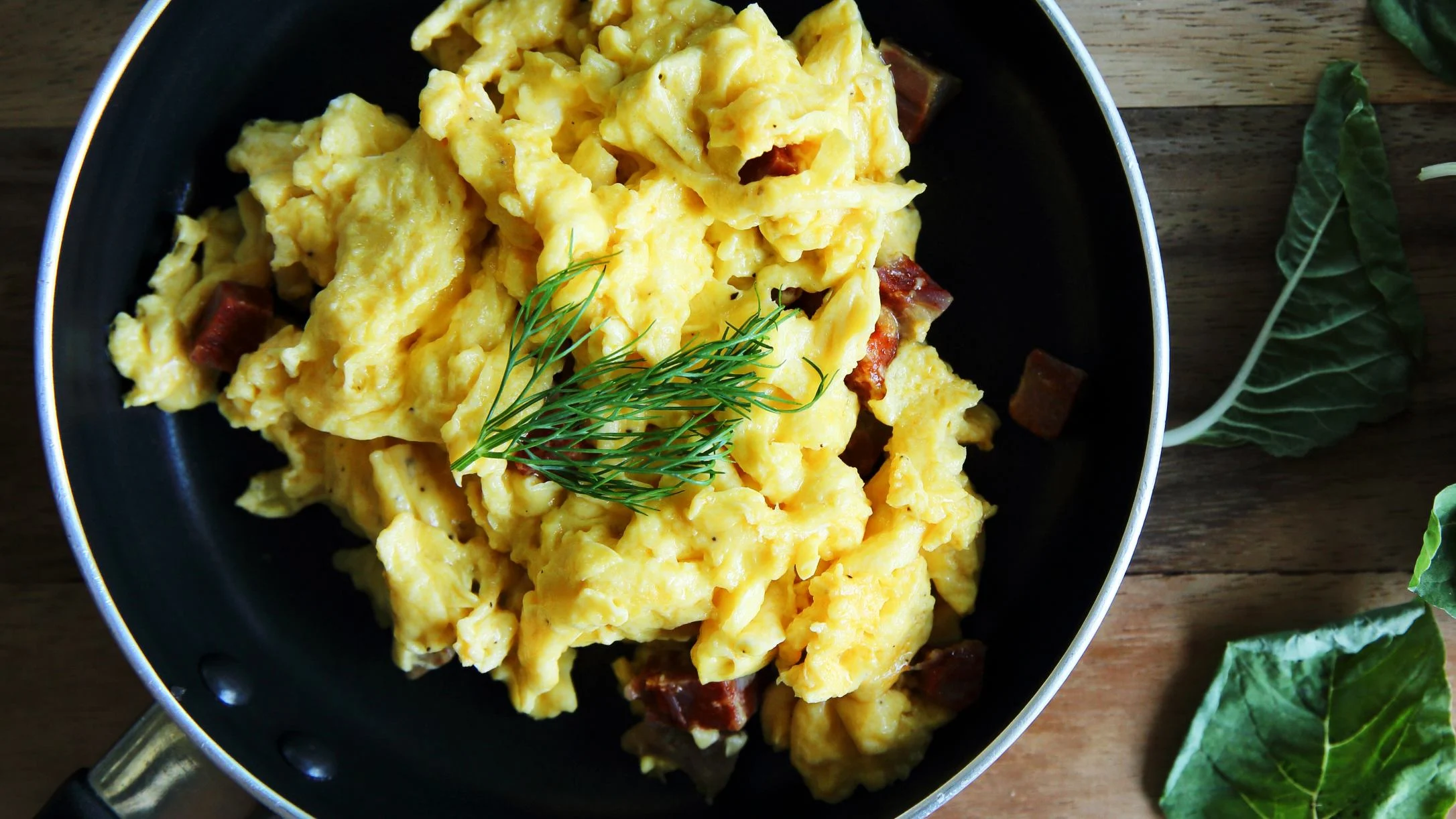 Fluffy yellow scrambled eggs with diced meat in a black skillet, garnished with fresh dill, on a wooden surface.