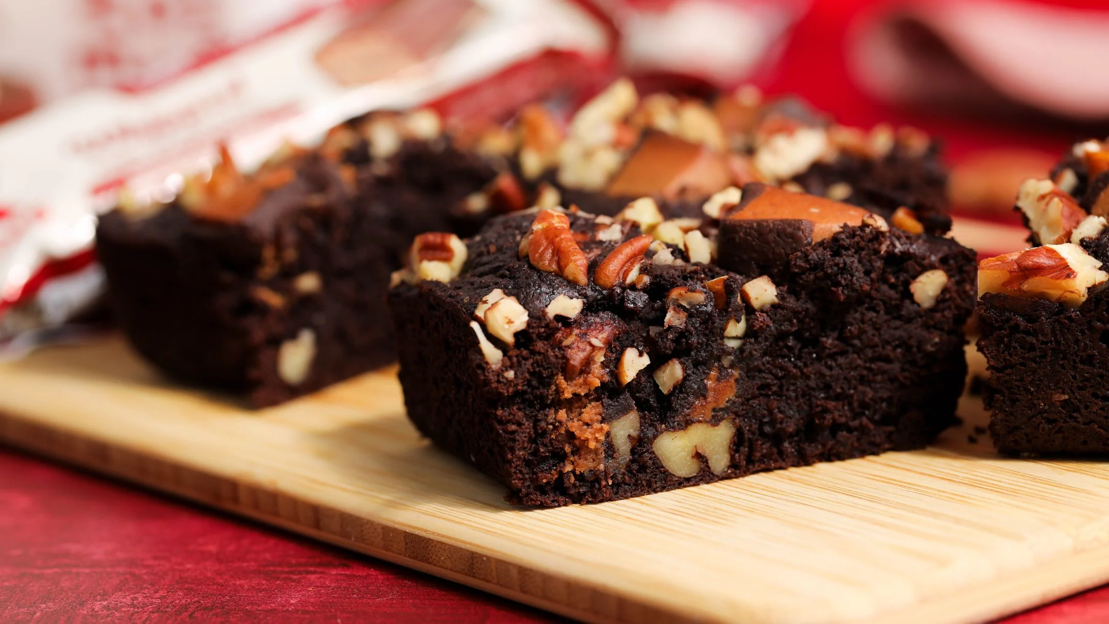 Fudgy chocolate brownies topped with chopped pecans on a wooden cutting board, with a red background and packaging behind.