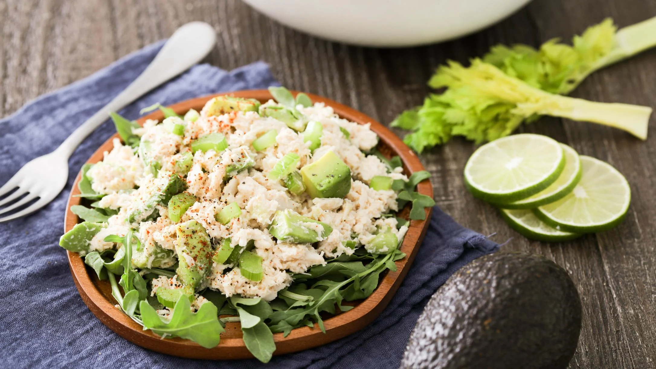 Crab salad with avocado and celery on arugula in a wooden bowl, with lime slices and celery stalks nearby.