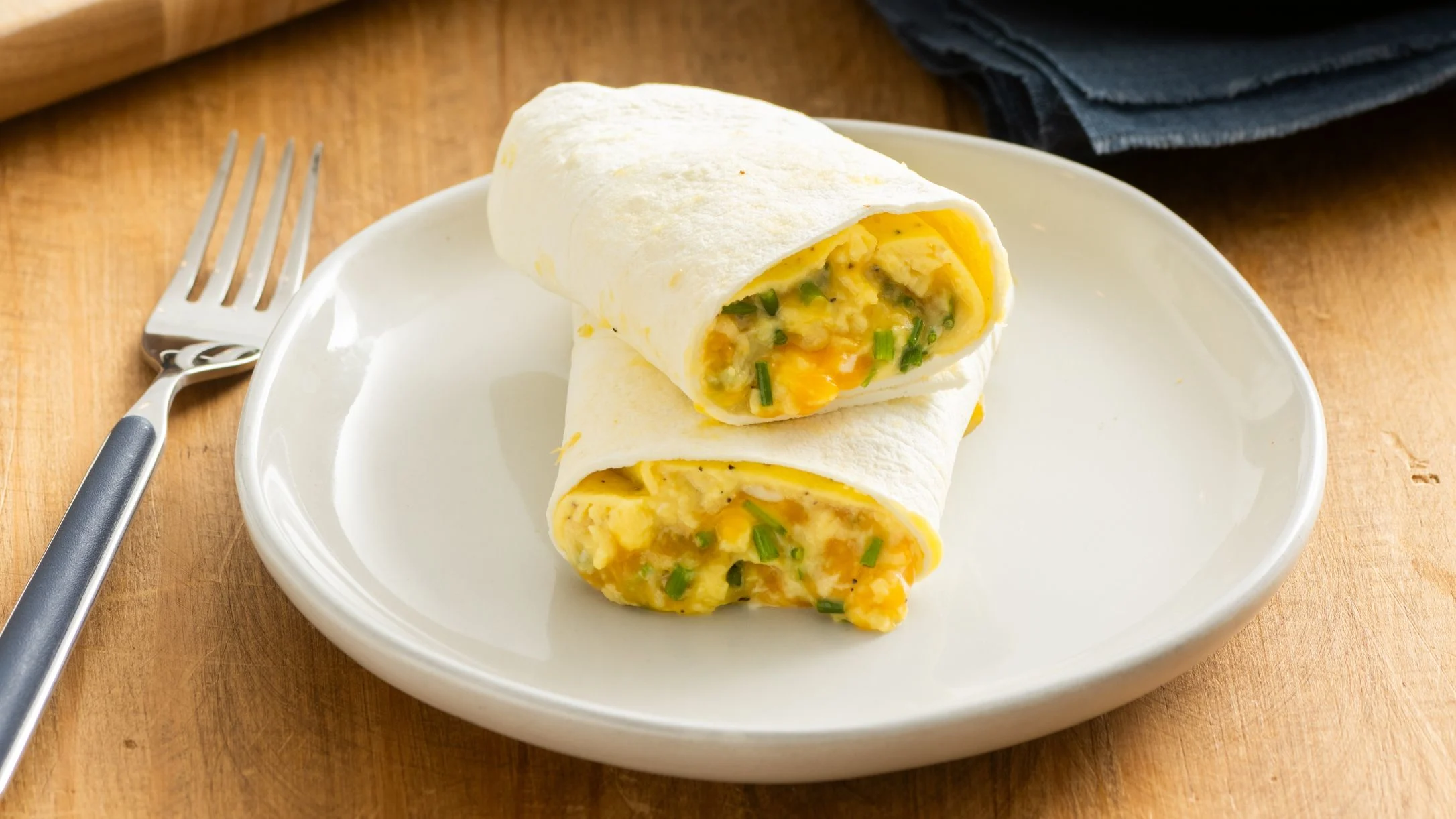 Breakfast burrito with scrambled eggs and green onions on a white plate, served on a wooden table with a fork.