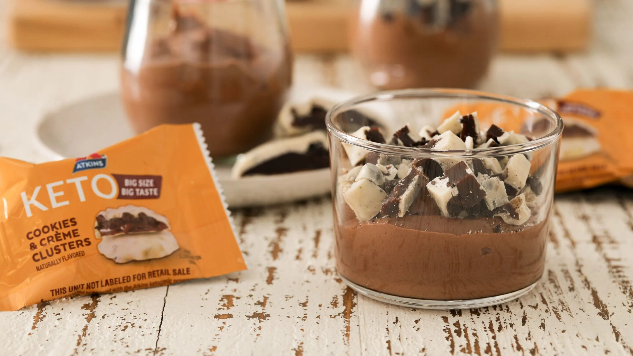 Chocolate pudding topped with cookie pieces next to Atkins Keto Cookies & Crème Clusters package on rustic wooden surface.