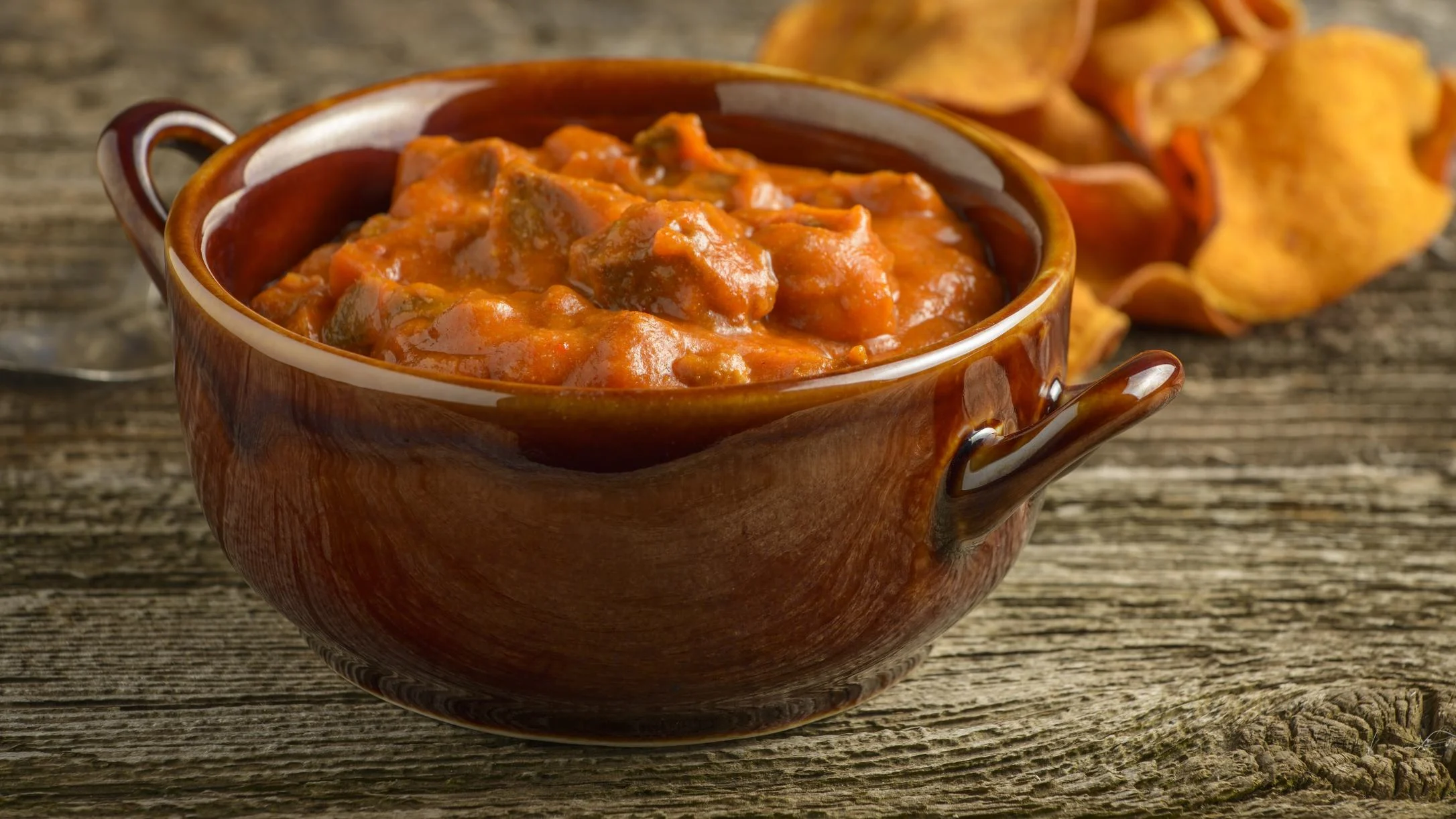 A brown ceramic bowl filled with rich, red-orange meat chili stew on a rustic wooden surface.
