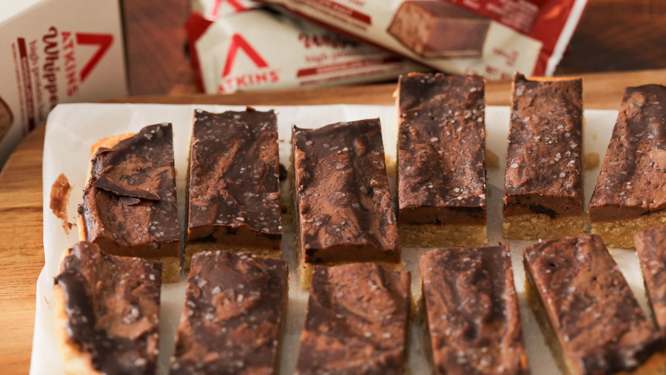 Chocolate-topped bar slices with sea salt on parchment paper, with Atkins product packaging visible in the background.