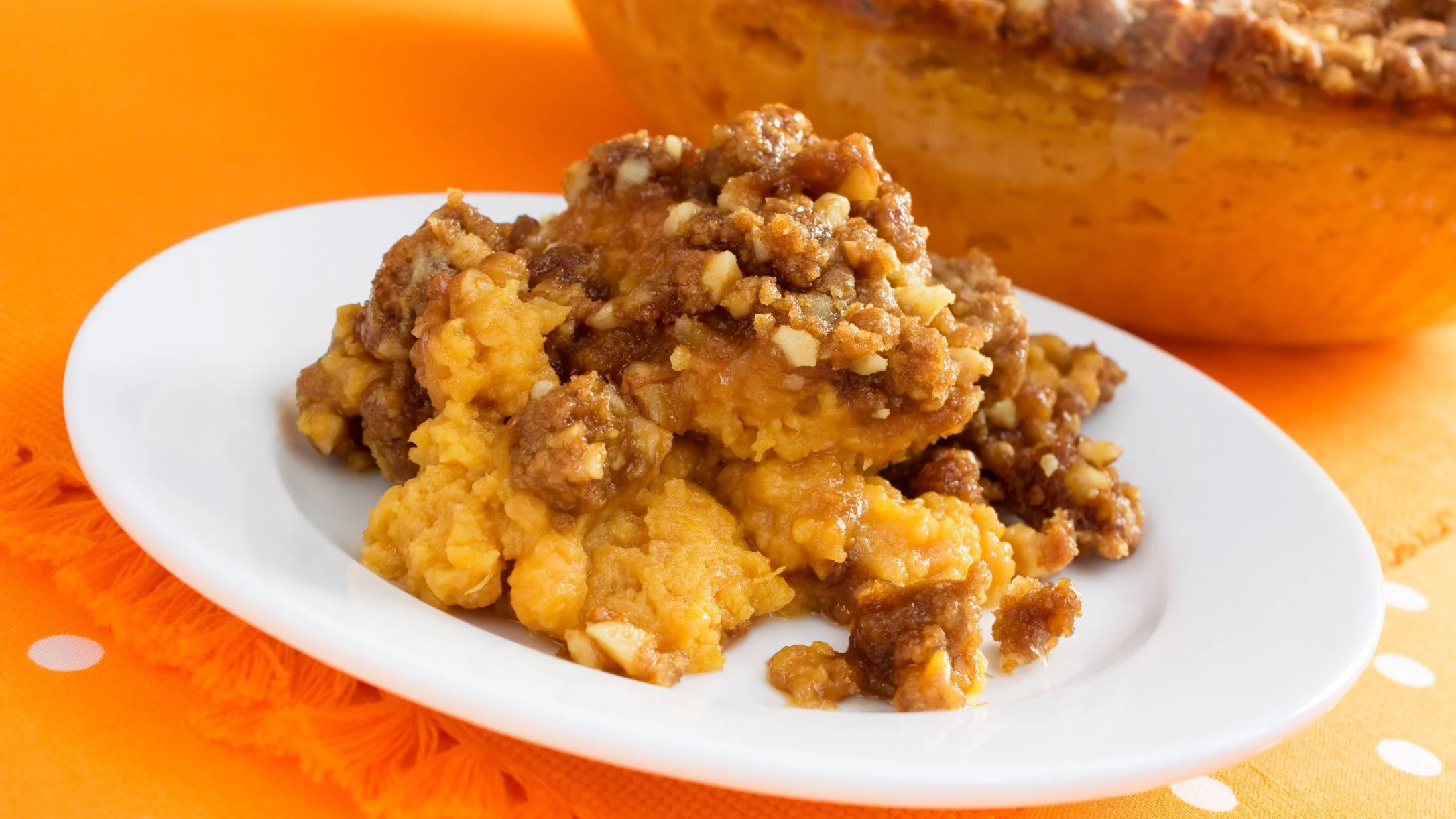 A serving of sweet potato casserole with a brown sugar pecan streusel topping on a white plate over an orange tablecloth.