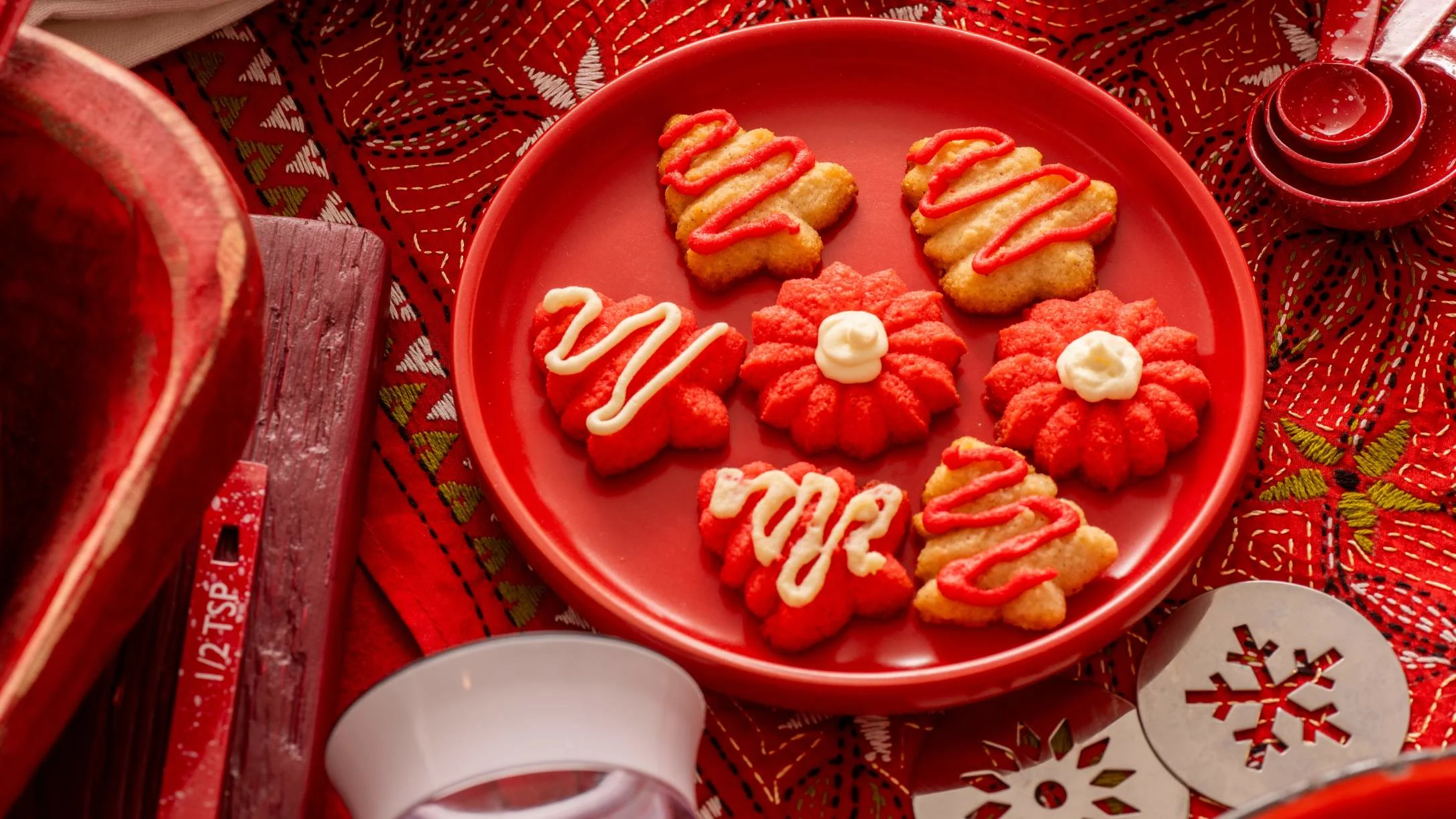 Red and yellow Christmas cookies with icing on a red plate, displayed on embroidered holiday fabric.