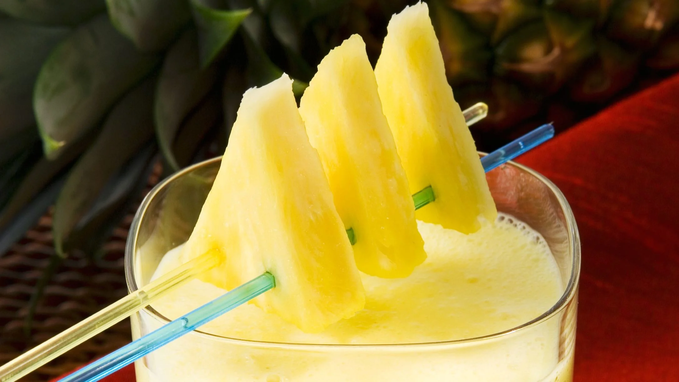 A creamy yellow pineapple smoothie in a glass, garnished with fresh pineapple slices and two colorful straws.