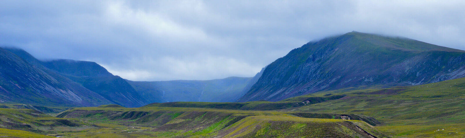 A cloudy sky over a valley surrounded by mountains in the Cairngorms National Park. A glen roves through the green landscape.
