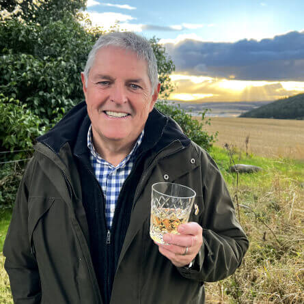 Scott Kelly, founder of Art of the Dram, holds a glass of Scotch Whisky in the garden with Highland scenery in the background.