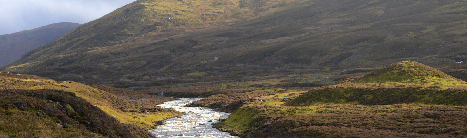 A river runs through a green valley in the Scottish Highlands whisky region. The sky is cloudy. Sun shines on the hills.