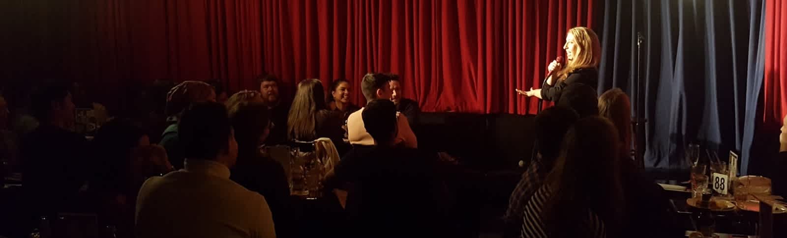 Pamela on stage doing standup in front of a red velvet curtain in a dimly lit packed room