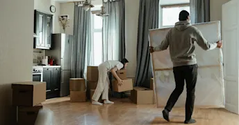par flyttar in i bostad Ett par flyttar in i bostad, kvinnan packar upp kartonger i köket och mannnen bär en stor tavla.