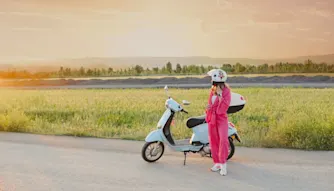 Helförsäkring moped Rosaklädd tjej står bredvid vit vespa längs vägen vid en äng.