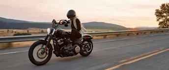 Person kör motorcykel på landsväg i solnedgång med skyddsutrustning och hjälm. En motorcyklist kör längs en landsväg i solnedgången, omgiven av öppna fält och berg i bakgrunden. Föraren bär hjälm, skyddsväst och mörka kläder och kör en svart custommotorcykel. Bilden fångar känslan av frihet, fart och sommarkvällar på två hjul – perfekt för artiklar om motorcykelkörkort, mc-försäkring eller livsstil med motorcykel.