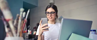 starta-enskild-firma.jpg En kvinna sitter omgiven av penslar och pennor vid sin dator med en telefon i handen