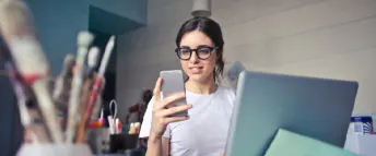 En kvinna sitter omgiven av penslar och pennor vid sin dator med en telefon i handen